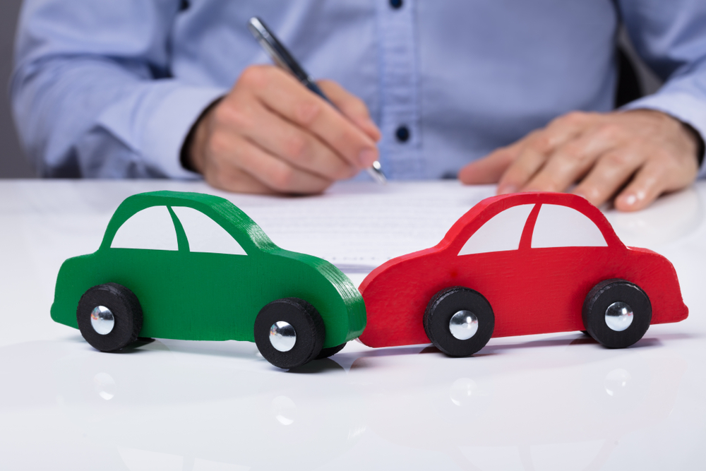 Two toy cars, one green and one red, positioned as if in a head-on collision on a white desk, with a person in a blue shirt signing a document in the background.