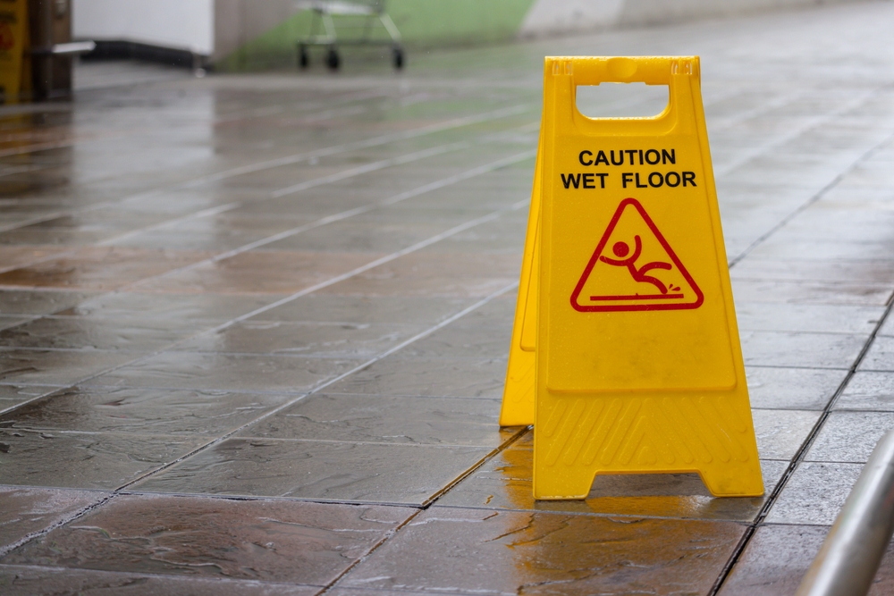 A yellow "Caution Wet Floor" sign standing on a wet, tiled indoor walkway.