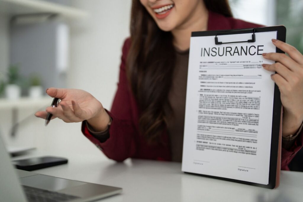 A smiling woman in a maroon blazer holding a clipboard with a document titled INSURANCE, gesturing toward a laptop on a white desk.